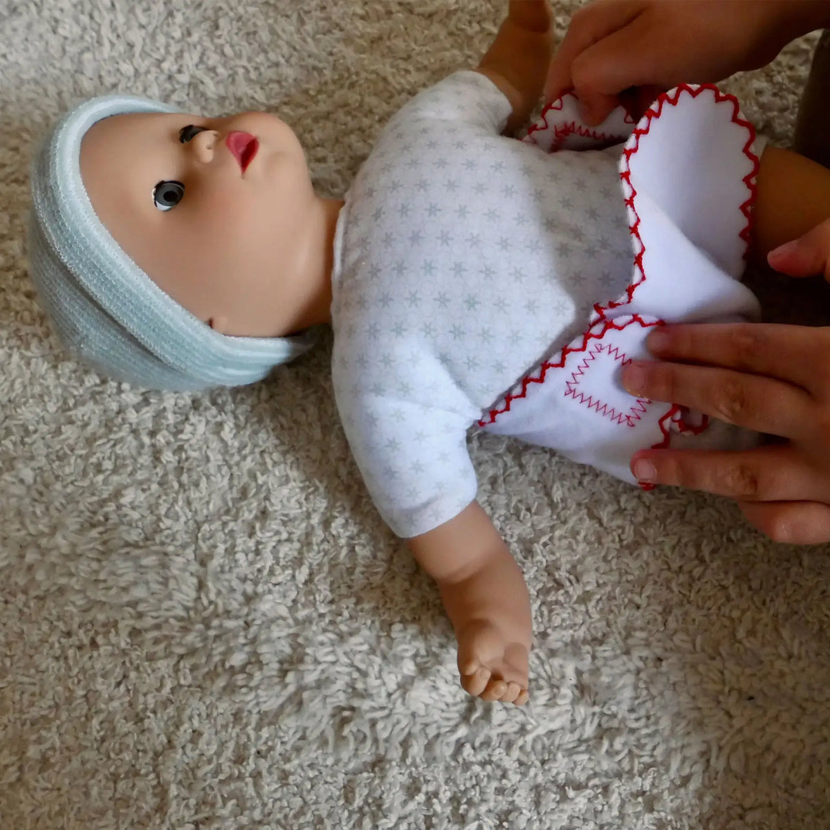 Götz Muffin To Dress Babypuppe wird von Kinderhänden angezogen, trägt weiße Hose mit roten Stickereien und weißes Hemdchen