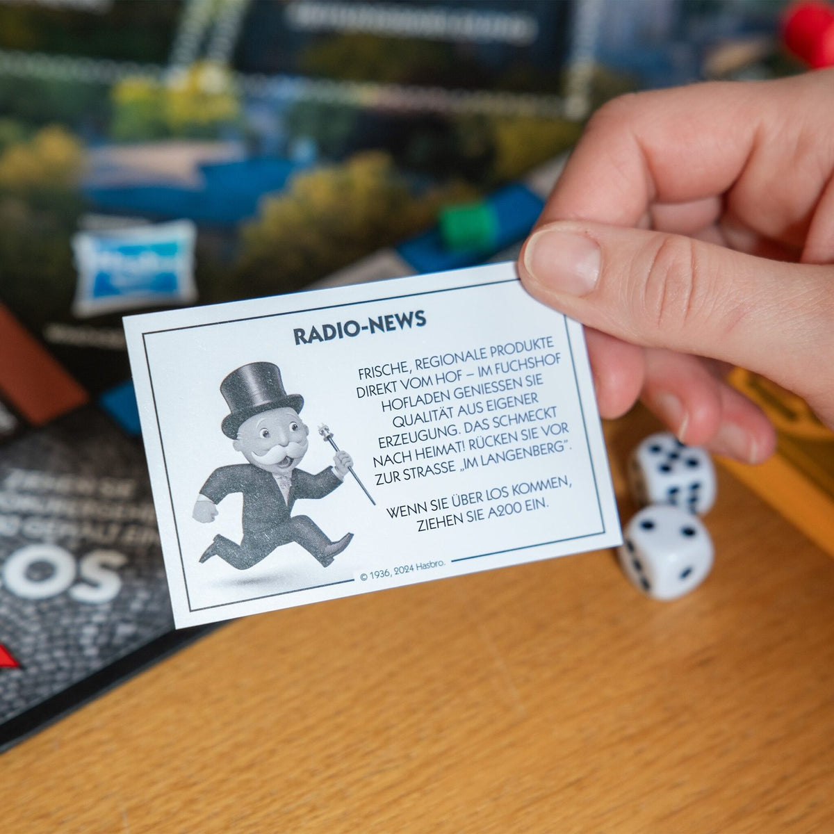 Hand hält eine Radio-News-Karte von Monopoly Konstanz, im Hintergrund Würfel und Spielbrett
