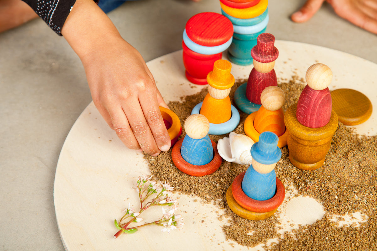 Kinderhand spielt mit Grapat Aguamarina Summer, bunte Holzfiguren mit Ringen auf weißer Platte mit Sand