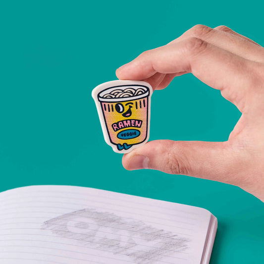 Hand hält OMY Eraser Buddies Noo Radierer als gelben Ramen-Becher vor türkisblauem Hintergrund