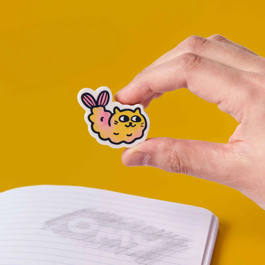 Hand hält den Radierer OMY Eraser Buddies Mewchi, eine Tempura-Garnele mit Katzengesicht, vor gelbem Hintergrund