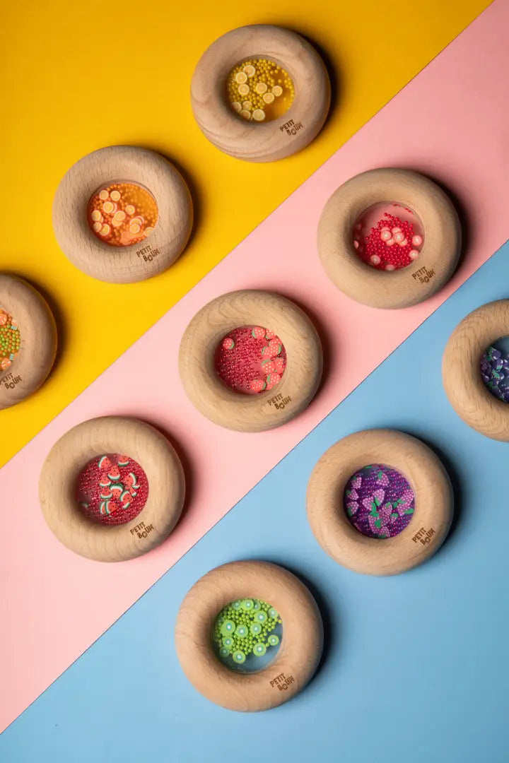 Mehrere Petit Boum Sensory Donuts mit verschiedenen Obst- und Gemüsefiguren auf gelb-rosa-blauem Hintergrund