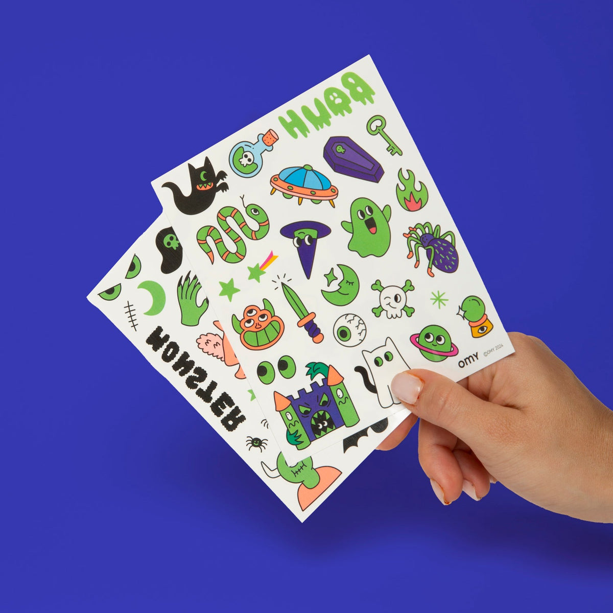 OMY Tattoos Boo Bogen mit bunten Halloween-Motiven wie Gespenster, UFO, Monster und Spinnen, Hand hält ihn vor blauem