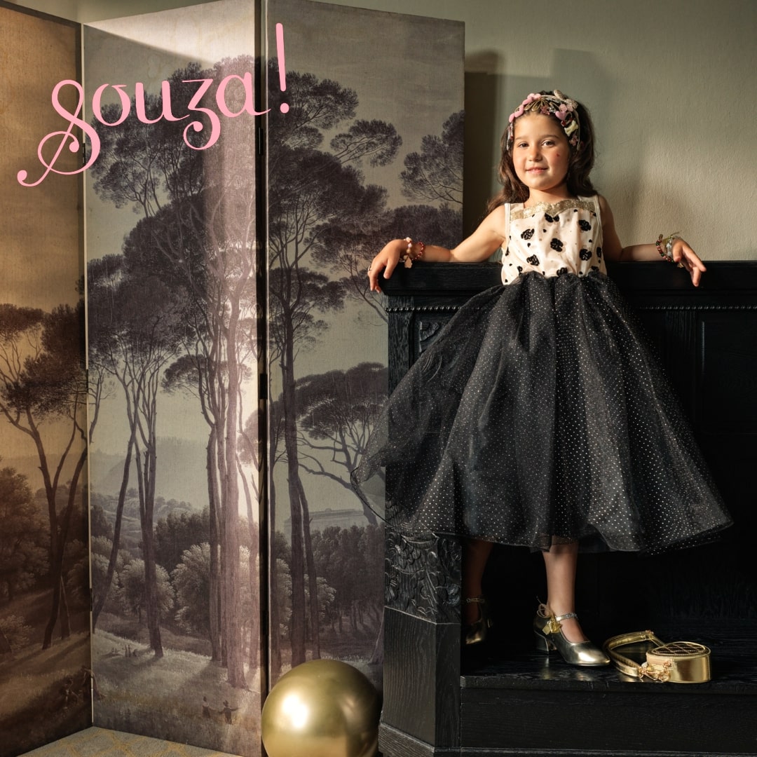 Kind trägt Souza Noella Kostümkleid mit cremefarbenem Oberteil und schwarzem gepunkteten Tüllrock, sitzt auf schwarzer Truhe