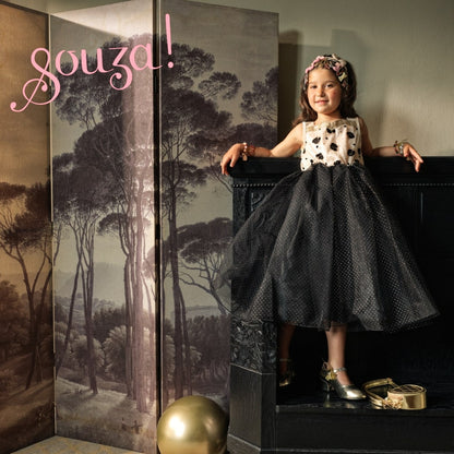 Kind trägt Souza Noella Kostümkleid mit cremefarbenem Oberteil und schwarzem gepunkteten Tüllrock, sitzt auf schwarzer Truhe