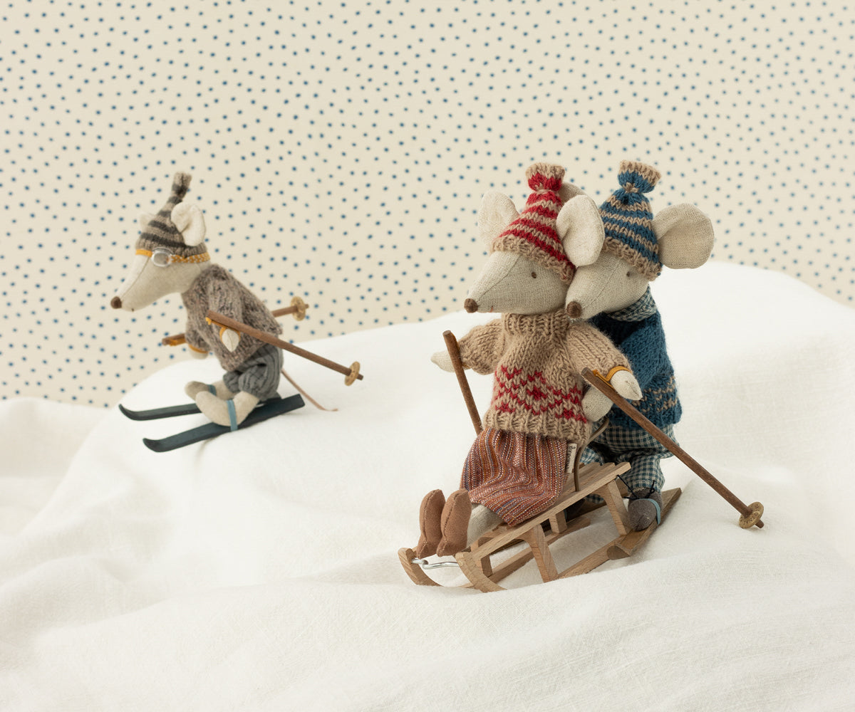 Zwei Maileg Mäuse mit Strickmützen fahren auf dem Holzschlitten durch den Schnee, daneben eine Maus auf Skiern