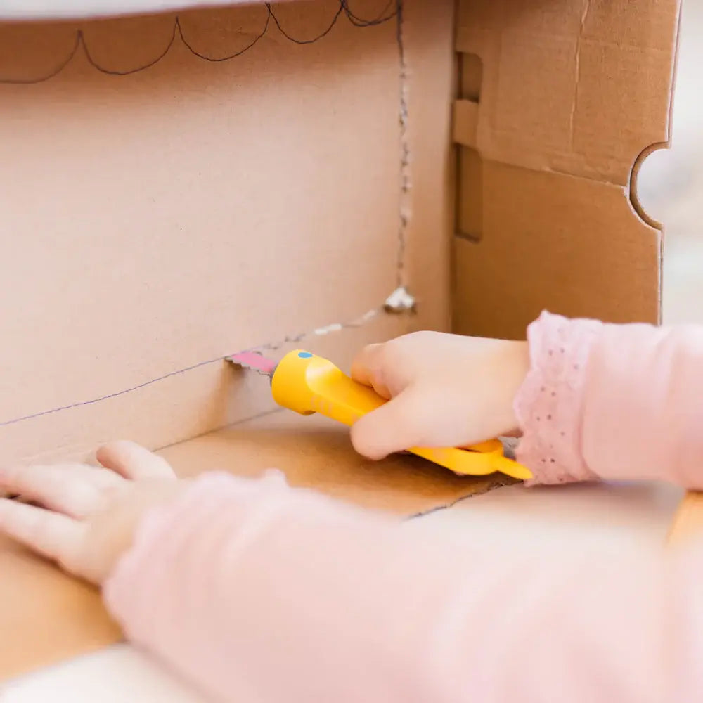 Kind in rosa Kleidung sägt mit gelber Makedo Safe-Saw Kartonsäge durch braune Pappschachtel, Detailaufnahme der Hände