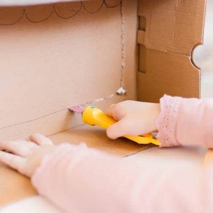 Kind in rosa Kleidung sägt mit gelber Makedo Safe-Saw Kartonsäge durch braune Pappschachtel, Detailaufnahme der Hände