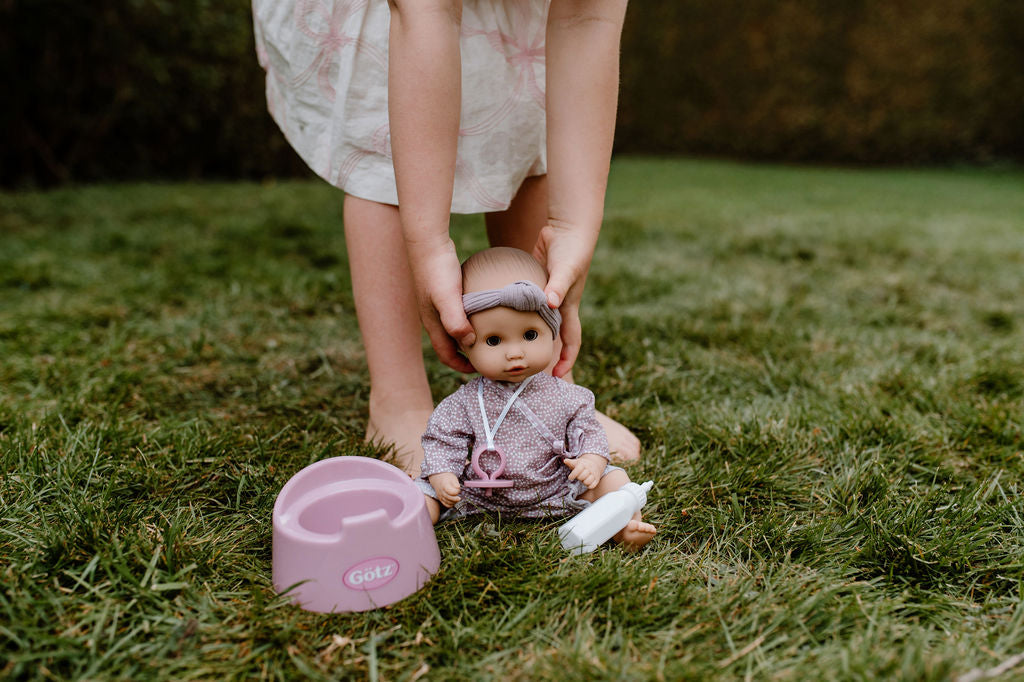 Kind hält die Götz Sleepy Aquini Girl Babypuppe in rosa Strampler und lila Stirnband auf grüner Wiese neben einem rosa Topf