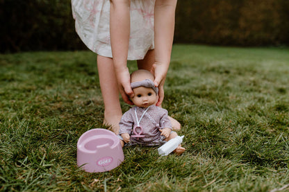 Kind hält die Götz Sleepy Aquini Girl Babypuppe in rosa Strampler und lila Stirnband auf grüner Wiese neben einem rosa Topf