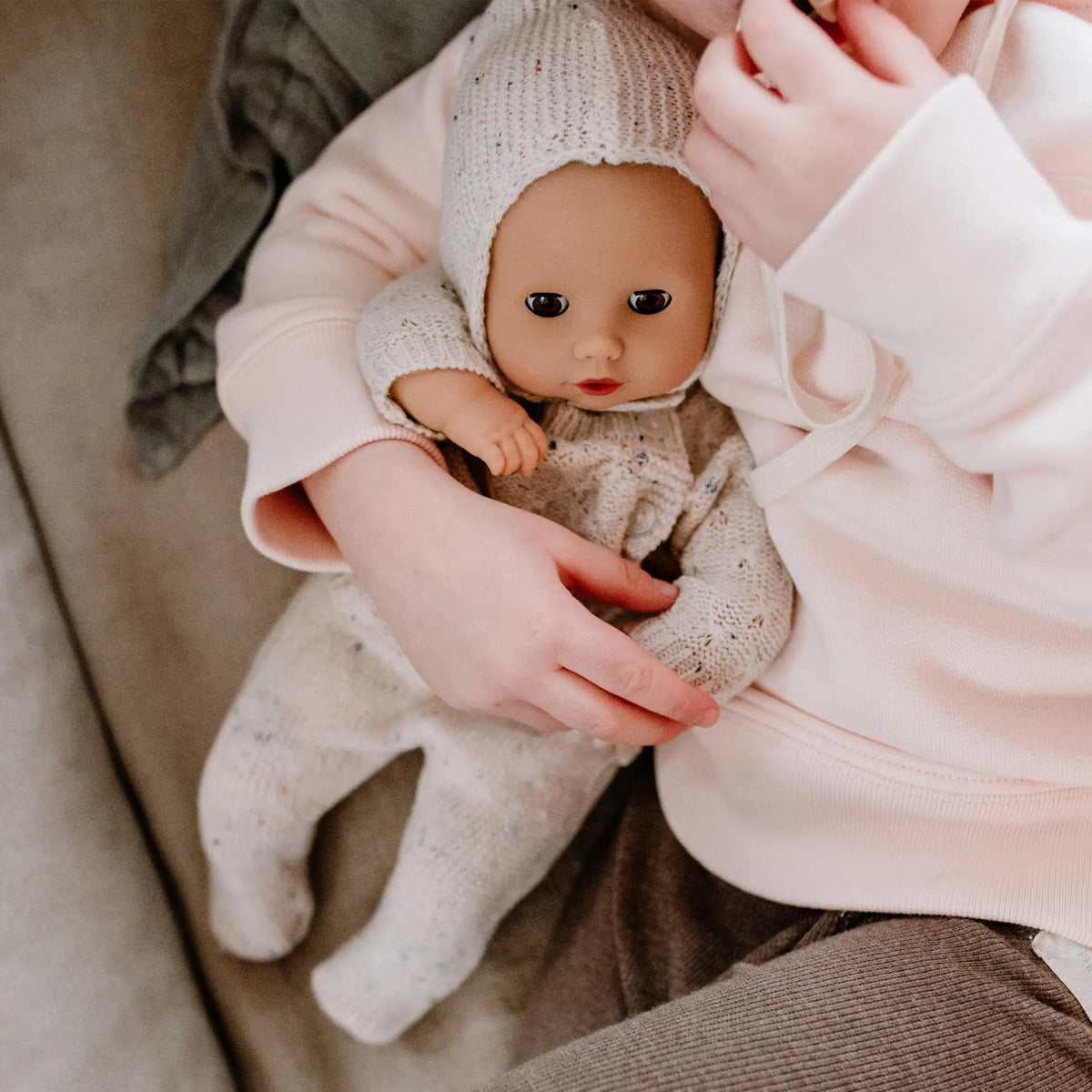 Götz Muffin Baby Alles Strick wird in beigem Strickanzug und Mütze von Kinderhänden in rosa Kleidung gehalten