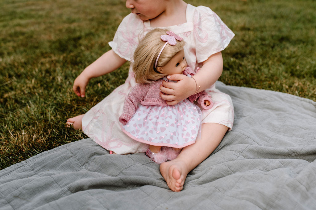 Kind sitzt mit Götz Muffin Puppe auf grauer Decke im Gras, hält die Puppe mit blondem Haar und rosa Kleid mit Herzchen-Muster