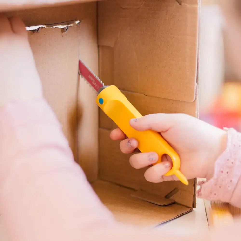 Kind sägt mit gelber Makedo Safe-Saw Kartonsäge durch braune Pappschachtel