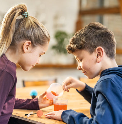 Zwei Kinder experimentieren mit dem Moses Kristallset, eines gießt orange Flüssigkeit in ein Glas