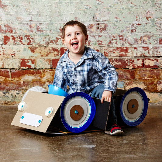 Lachendes Kind sitzt in einem mit Makedo gebauten Karton-Auto mit großen blauen Rädern