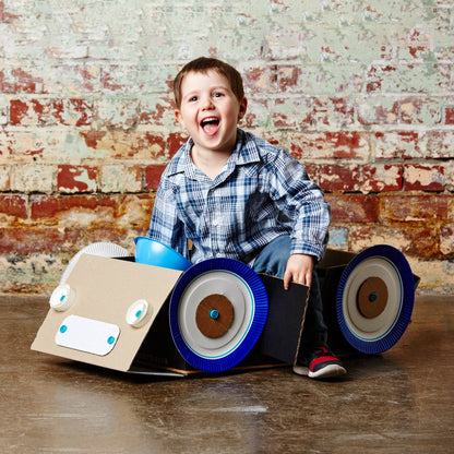 Lachendes Kind sitzt in einem mit Makedo gebauten Karton-Auto mit großen blauen Rädern