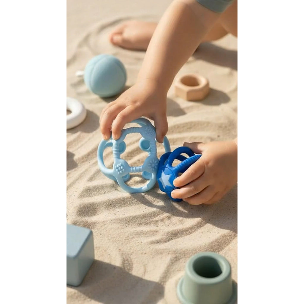 Kind greift im Sand nach den beiden blauen Sensorikbällen, im Hintergrund Stapelteile aus Silikon