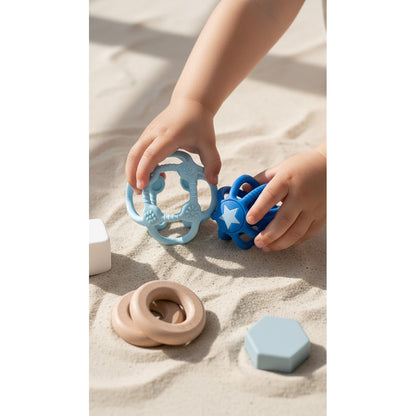 Kind spielt im Sand mit beiden blauen Sensorikbällen, daneben beige Silikonringe