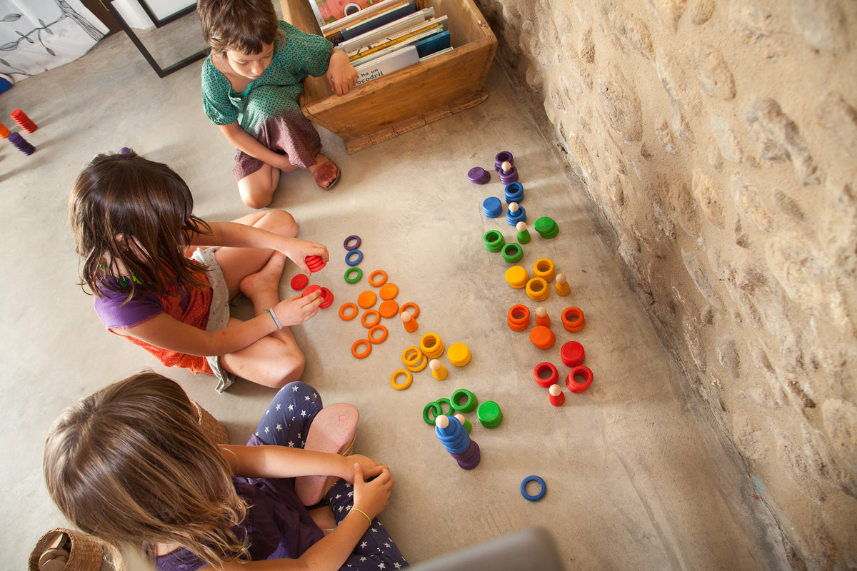 Drei Kinder spielen auf hellem Boden mit Grapat Nins Rings, bunte Ringe verteilt zu verschiedenen Mustern arrangiert