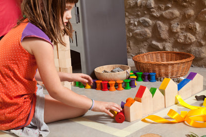 Kind in orangem Shirt spielt mit Grapat Houses and Nins, arrangiert bunte Holzbausteine und Häuser auf weißer Spielfläche