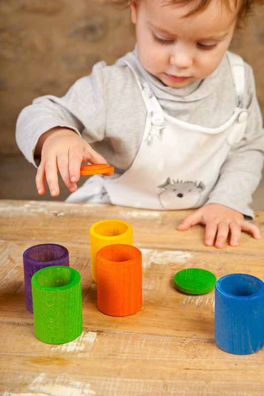 Kleines Kind beim Spielen mit Grapat Farbbechern in Violett, Grün, Orange, Gelb, Blau und deren Deckeln auf Holztisch