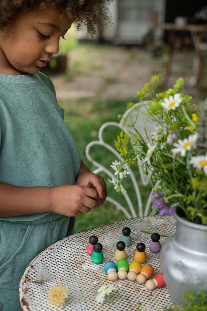 Kind spielt mit Grapat Dark Baby Nins Holzfiguren an Tisch im Garten neben Blumentöpfen mit gelben und weißen Blüten
