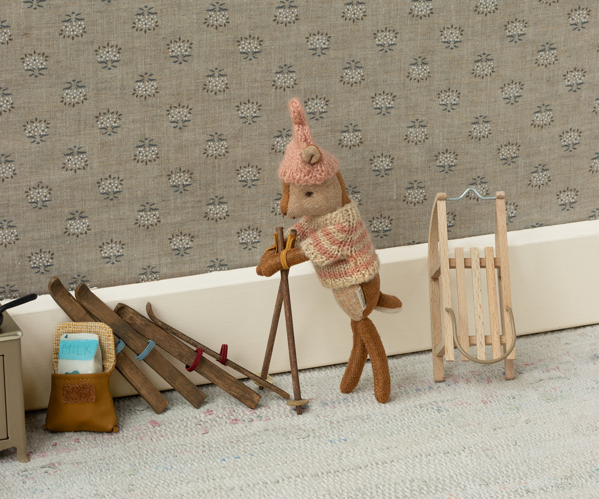 Maileg Holzschlitten lehnt an der Wand neben einer Figur mit Skistöcken und mehreren Paar Skiern