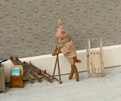 Maileg Holzschlitten lehnt an der Wand neben einer Figur mit Skistöcken und mehreren Paar Skiern