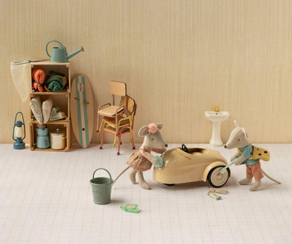 Maileg Fahrerin-Maus und eine zweite Maus putzen ein cremefarbenes Mausauto, dahinter Regal und Stühle
