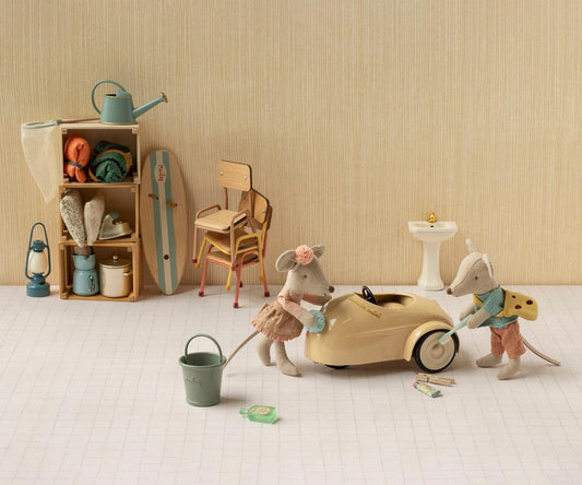 Maileg Fahrerin-Maus und eine zweite Maus putzen ein cremefarbenes Mausauto, dahinter Regal und Stühle
