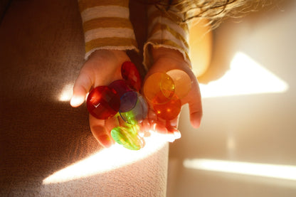 Kinderhände halten rote, orange und gelbgrüne Funkelsteine gegen Licht, die Farben leuchten durch