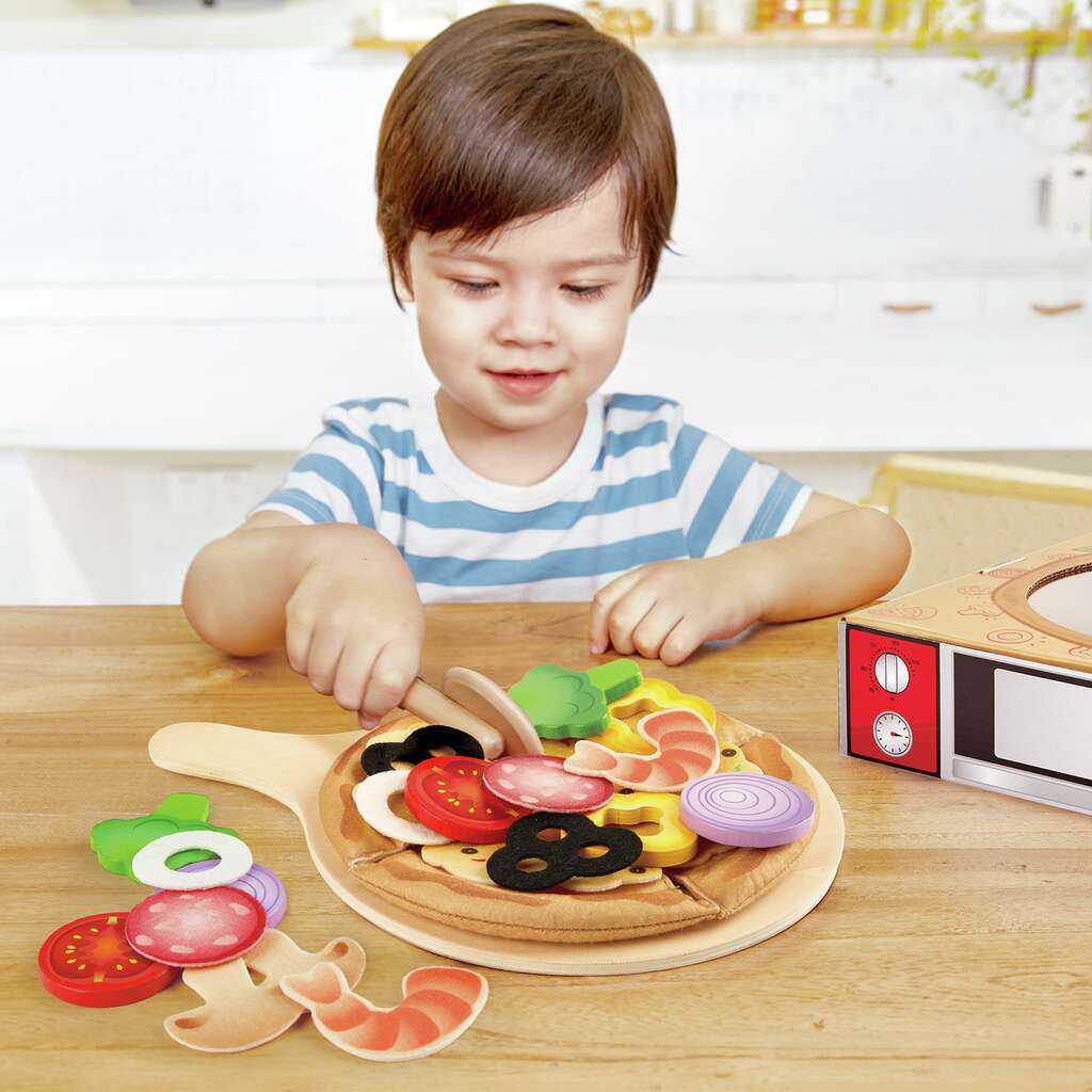 Kleines Kind in blau-weiß gestreiftem Shirt legt Holzgemüse auf Holzpizza beim Spielen mit dem Hape Pizza-Set