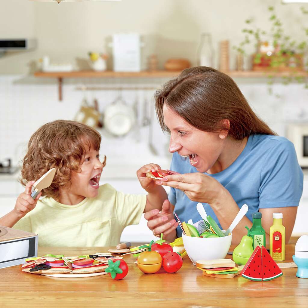 Frau und Kind spielen mit dem Hape Pizza-Set an einem Holztisch, beide lachen und halten Holzgemüseteile in den Händen