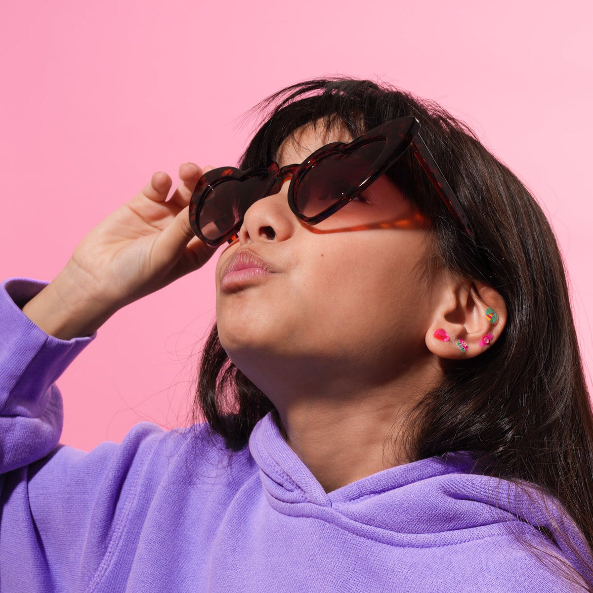 Kind trägt schwarze Sonnenbrille und bunte OMY Lily Ohrringe zum Aufkleben auf rosa Hintergrund