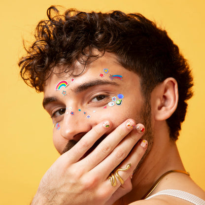 OMY Gesichtssticker Kawaii, bunte glitzernde Motive auf Stirn und Wange eines Mannes vor gelbem Hintergrund