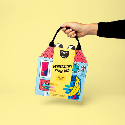 Hand hält Verpackung des OMY Montessori Play Kit Mein Tag mit rosa Dach und farbigen Illustrationen