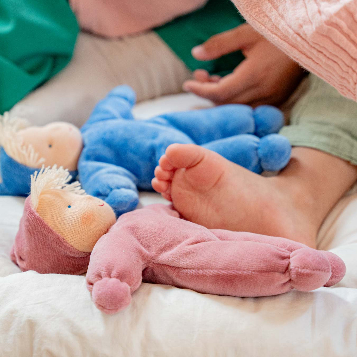 Zwei Käthe Kruse Waldorfpuppen LOLO in Altrosa und Blau liegen neben einem Baby auf einer Decke