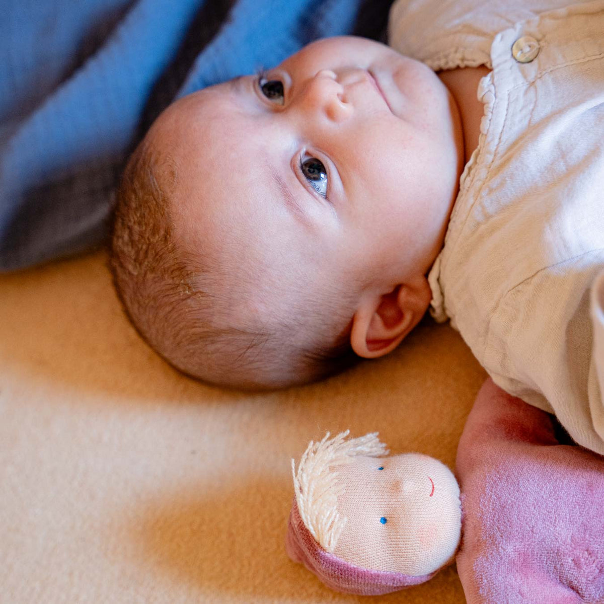 Baby liegt auf einer Decke und schaut nach oben, neben ihm die Käthe Kruse Waldorfpuppe LOLO rose
