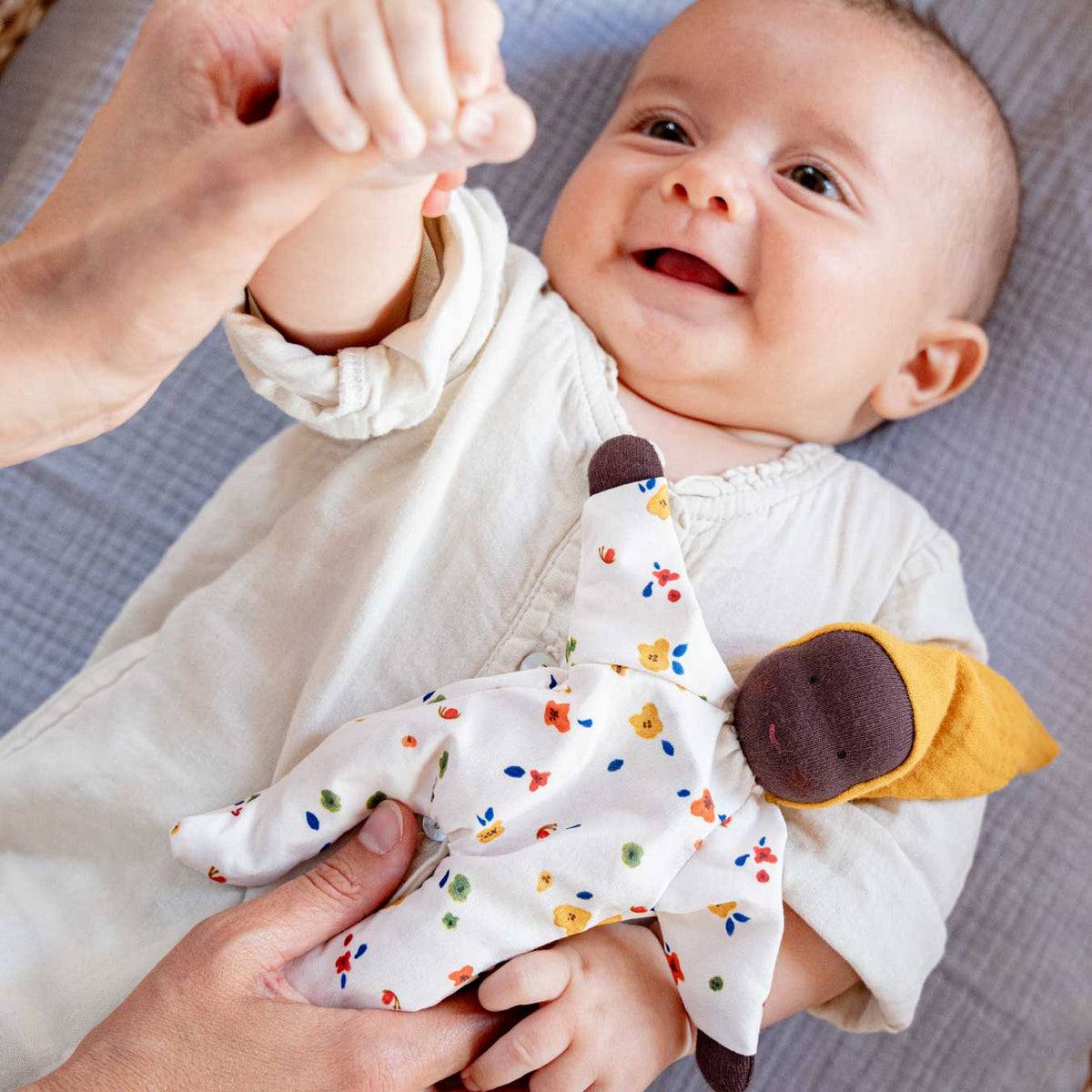 Lachendes Baby liegt auf einer Decke, auf seinem Bauch die Käthe Kruse Waldorfpuppe NINI Sonnenschein