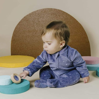 Kleinkind in blauem Strampler greift nach einem Reflect Donut aus Holz auf einem türkisen Podest