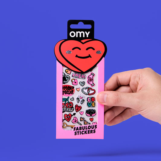 OMY Sticker-Packung mit rotem Herz-Anhänger, rosa Karton, Hand hält sie vor blauem Hintergrund