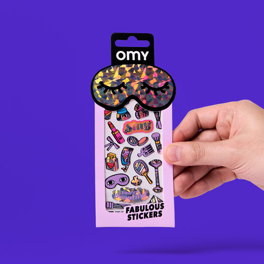 OMY Iconic Make-Up Sticker, Kind hält die rosa Verpackung mit Augenmaske-Motiv und bunten Sticker-Abbildungen
