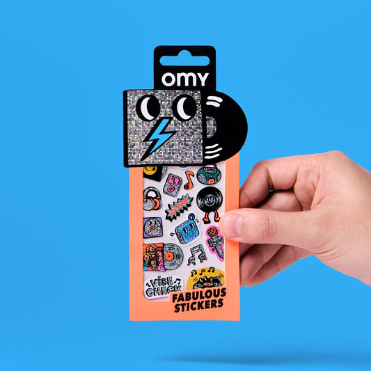 Hand hält OMY Iconic Musik-Sticker Packung mit bunten Motiven vor blauem Hintergrund