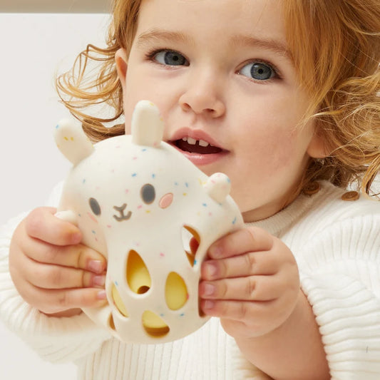 Kind mit rötlichen Locken hält die Tiger Tribe Silikon-Rassel Hase in Creme-Weiß mit goldgelben Löchern in beiden Händen