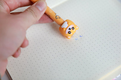 Hand hält den Stift mit Figur Shiba-Hund und radiert damit Schrift auf Punktrasterpapier
