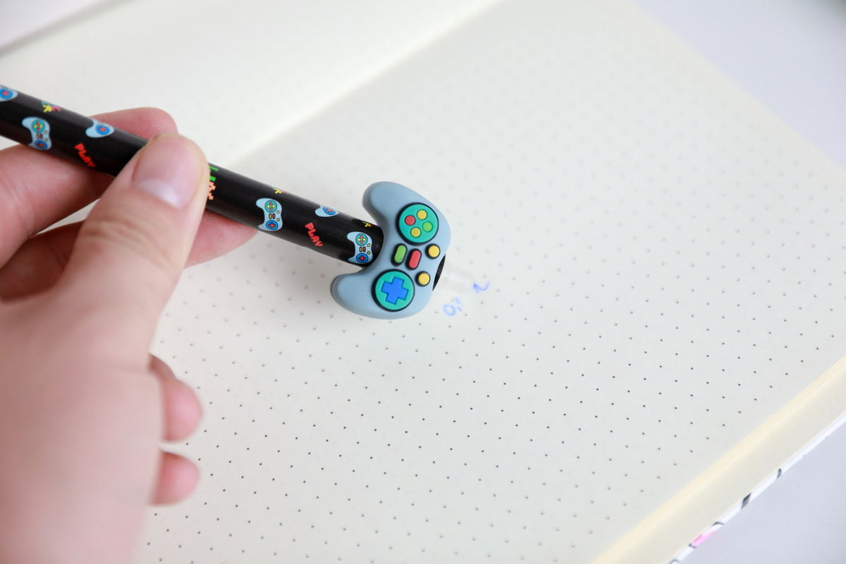 Hand hält den Stift mit Figur Gamecontroller und radiert damit Schrift auf Punktrasterpapier