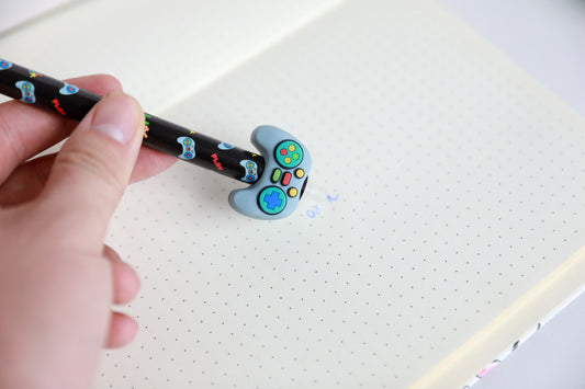 Hand hält den Stift mit Figur Gamecontroller und radiert damit Schrift auf Punktrasterpapier
