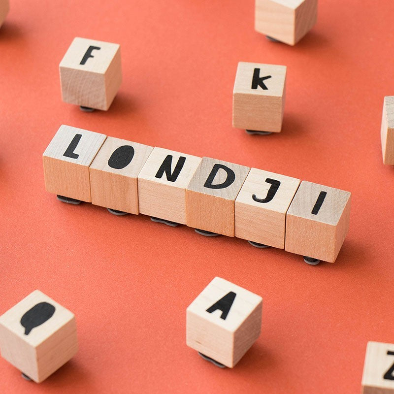 Holzwürfel mit Buchstabenstempeln, waagerecht das Wort LONDJI, einzelne Würfel mit f, k, a verstreut auf orangefarbenem