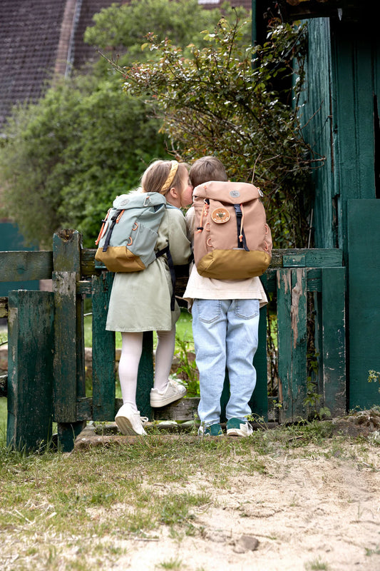 Zwei Kinder von hinten mit Lässig Wanderrucksäcken in Rosé und Grau beim Betreten eines grünen Gartentores