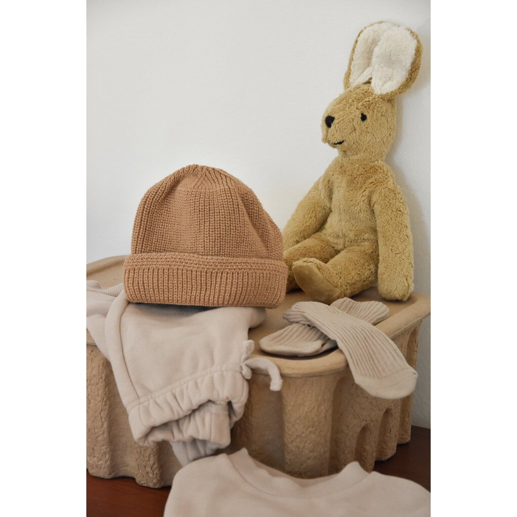 Senger Naturwelt Schlenker-Hase in Beige sitzt neben einer Strickmütze und Kleidung auf einem Holzhocker